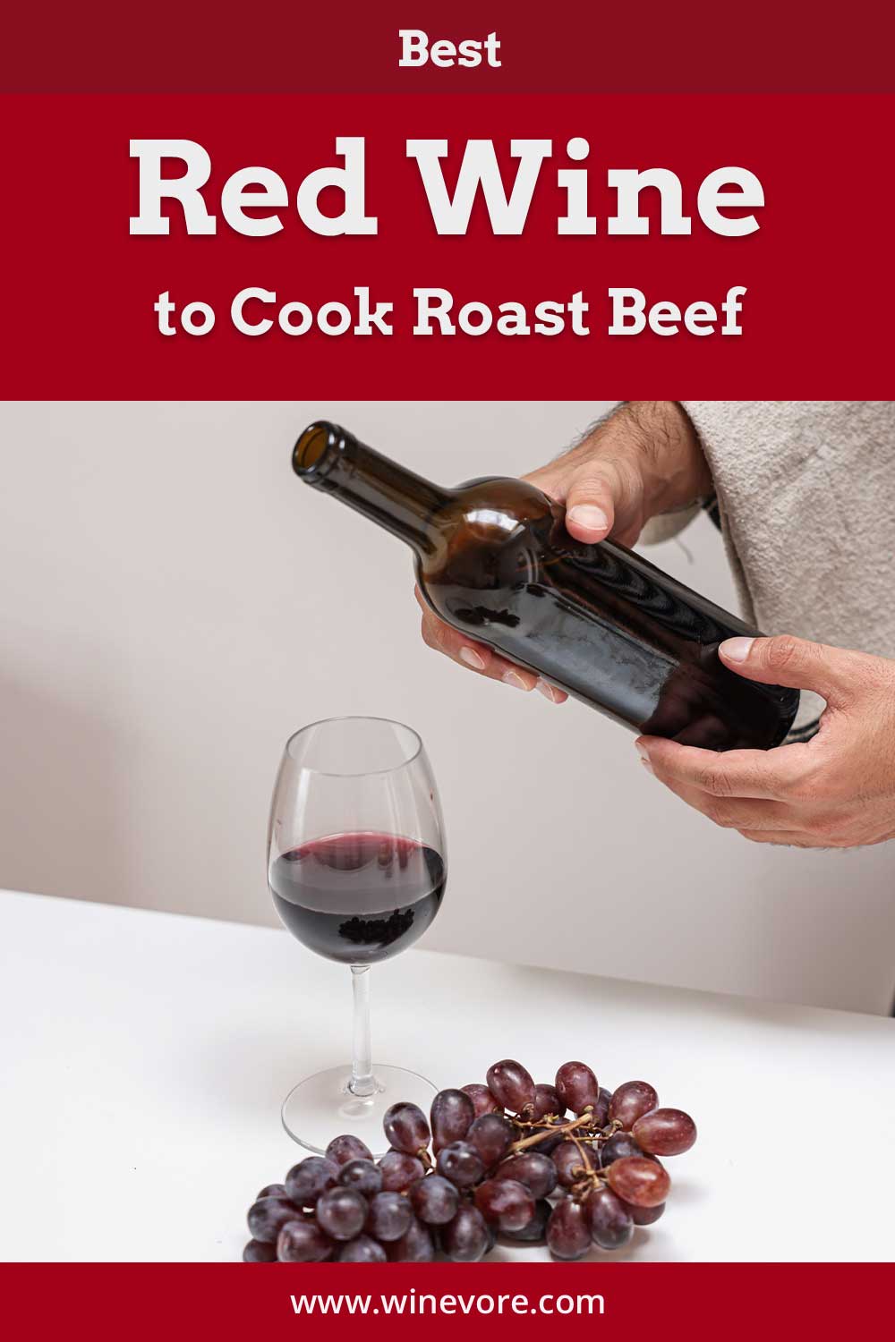A bottle of wine held by two hands over a glass of wine with a bunch of grapes on a table - Best Red Wine to Cook Roast Beef.